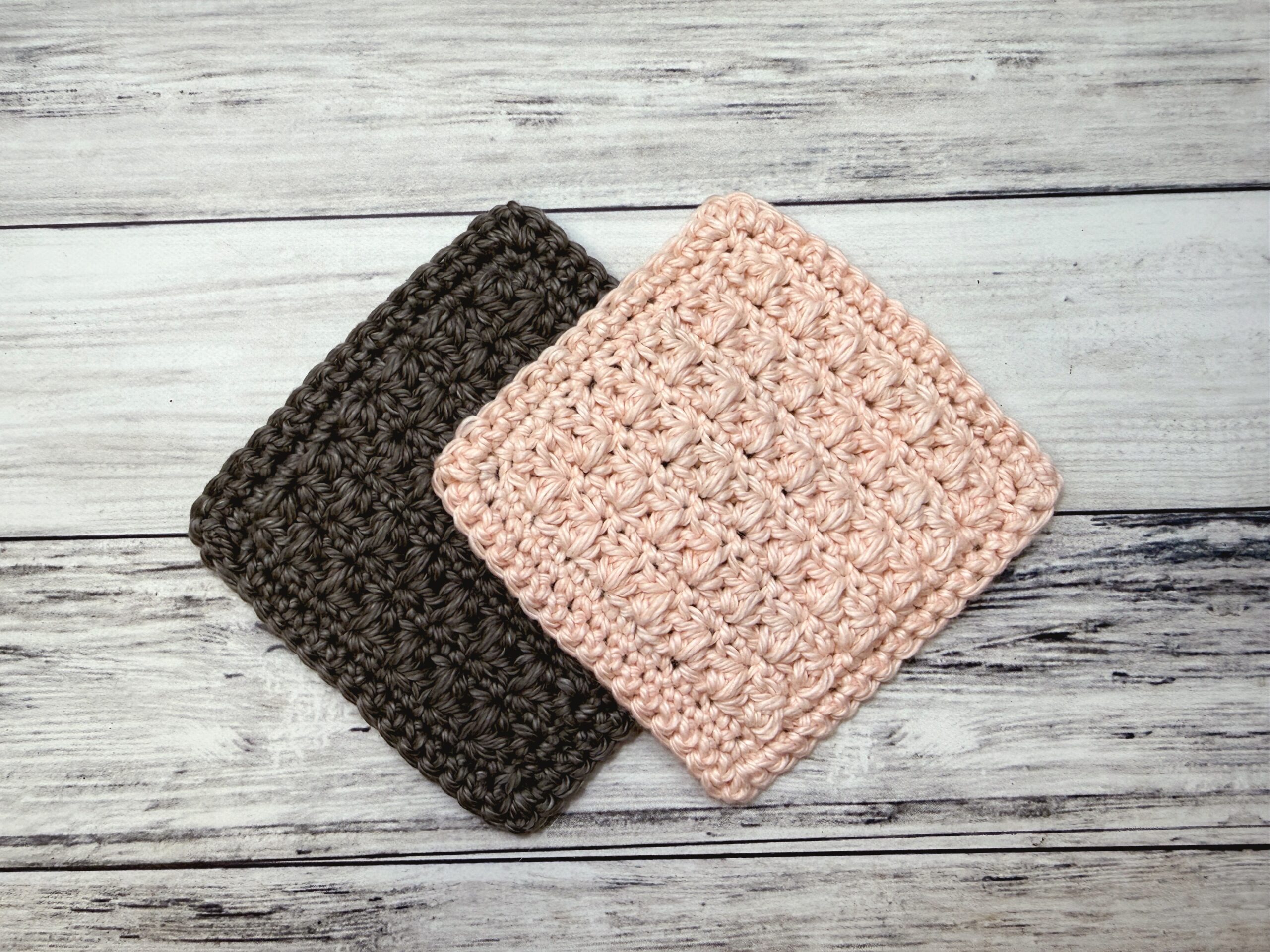 Two square crocheted cotton face scrubbies in textured cluster stitch, one pale pink and one charcoal gray, on white wood
